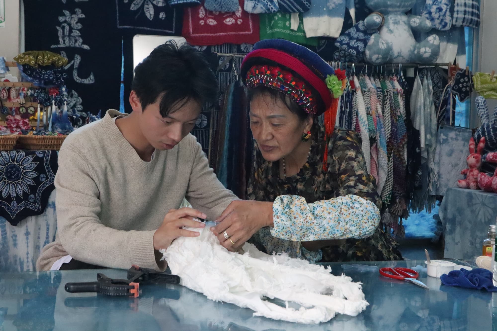 The grandmother guiding a young visitor's stitches at the workshop table